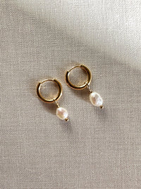 A pair of gold hoop earrings with dangling freshwater pearls, displayed flat on textured cream fabric.