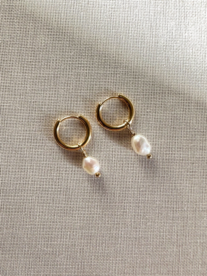 A pair of gold hoop earrings with dangling freshwater pearls, displayed flat on textured cream fabric.