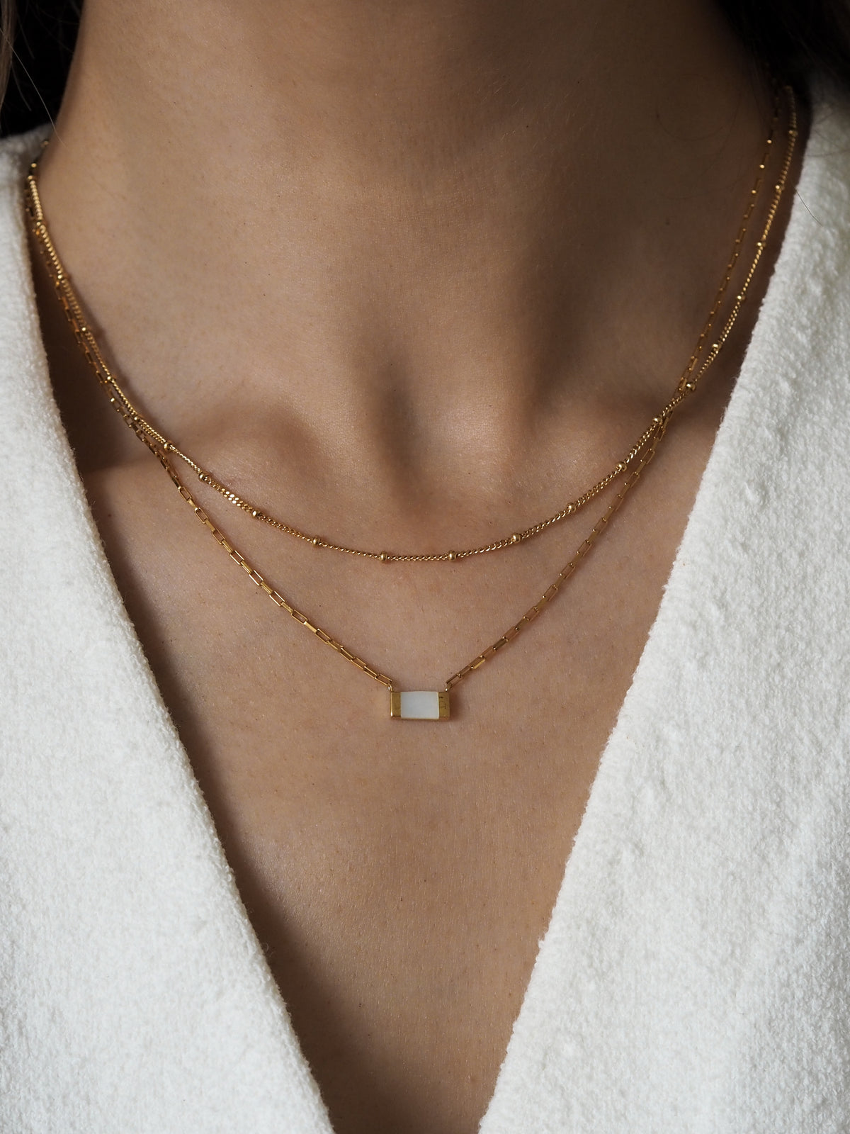 Layered gold necklaces worn on model, featuring a gold box chain necklace with a rectangular mother of pearl pendant and a delicate satellite chain. Styled with a white textured fabric top.