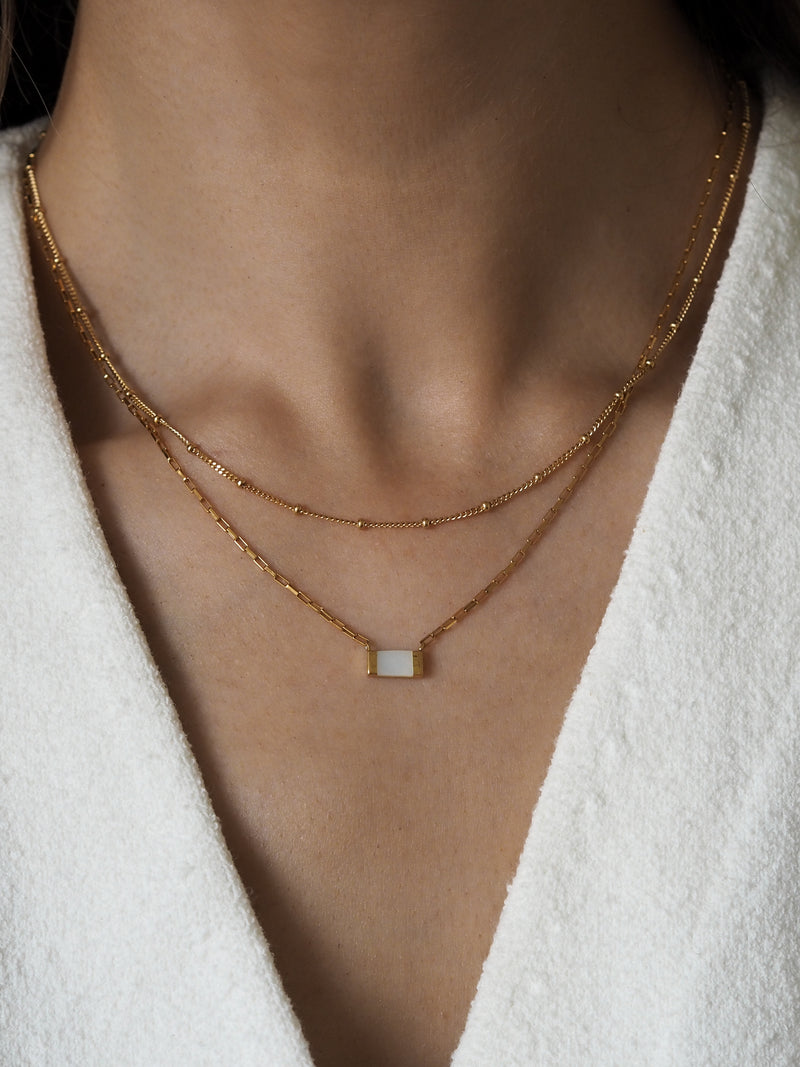 Layered gold necklaces worn on model, featuring a gold box chain necklace with a rectangular mother of pearl pendant and a delicate satellite chain. Styled with a white textured fabric top.