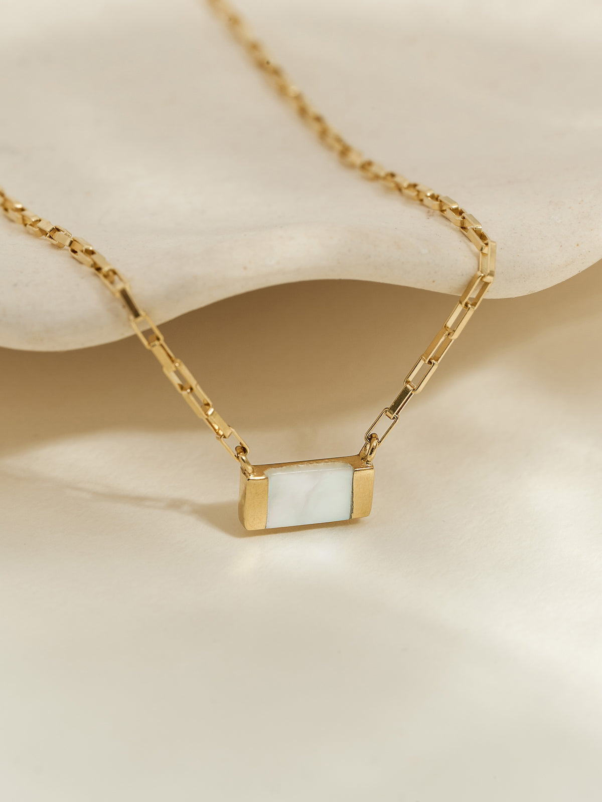 Close-up of a gold pendant necklace with a rectangular mother of pearl stone, displayed on a neutral textured surface.