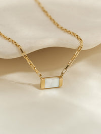 Close-up of a gold pendant necklace with a rectangular mother of pearl stone, displayed on a neutral textured surface.