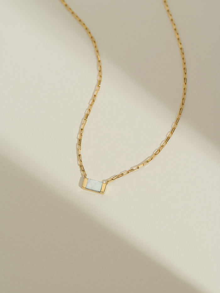Gold chain necklace with a rectangular mother of pearl pendant, photographed flat against a soft cream background with natural shadows.