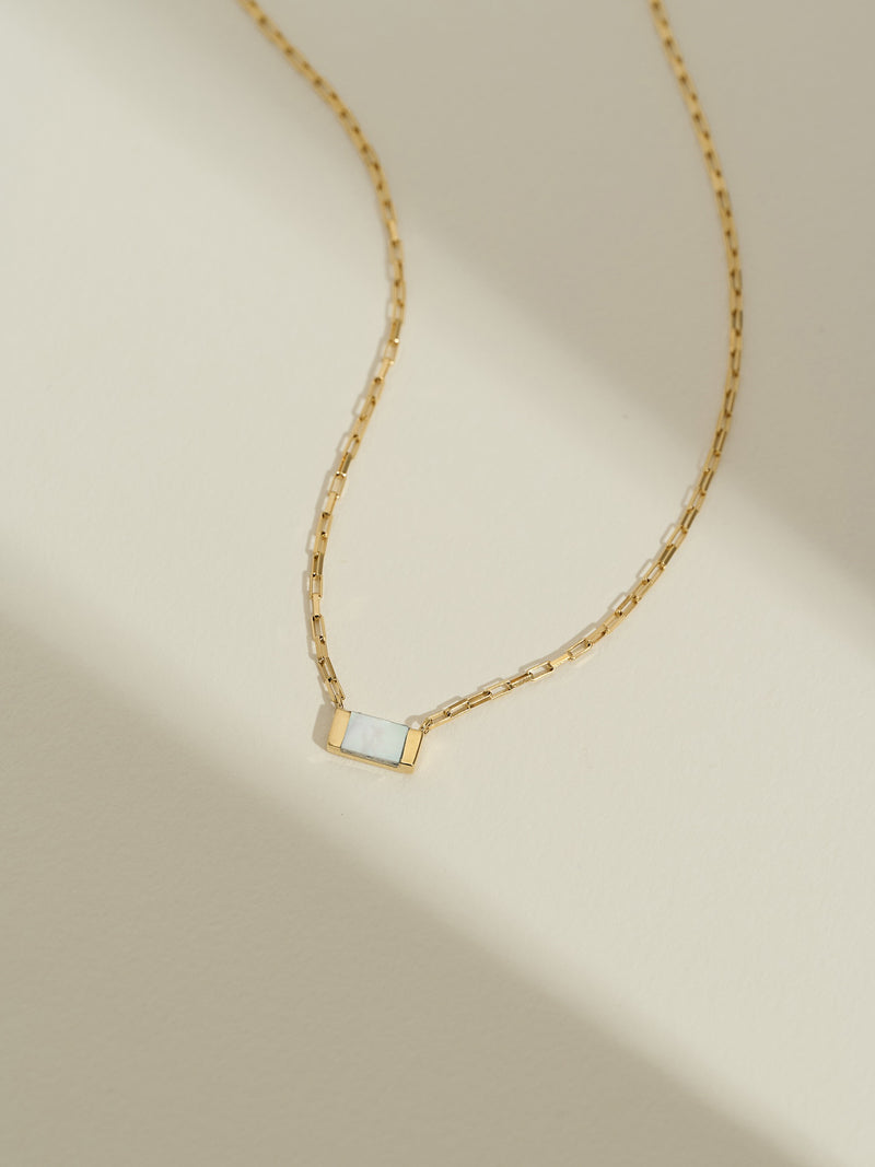 Gold chain necklace with a rectangular mother of pearl pendant, photographed flat against a soft cream background with natural shadows.