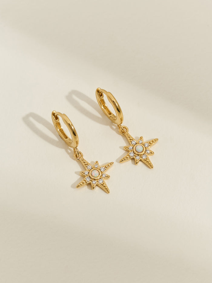 A pair of gold-plated huggie earrings with star-shaped drop charms featuring central opals and surrounding cubic zirconia stones, displayed on a cream background.