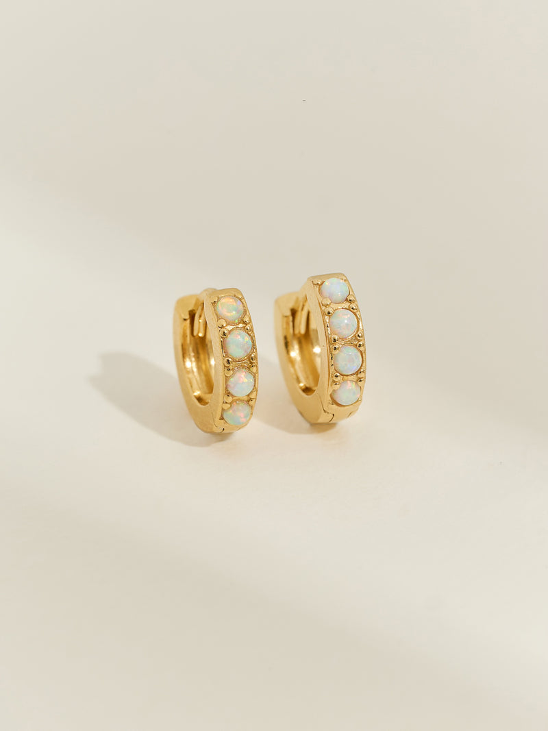 Pair of gold-plated huggie earrings set with five iridescent opal stones, photographed against a soft cream background.