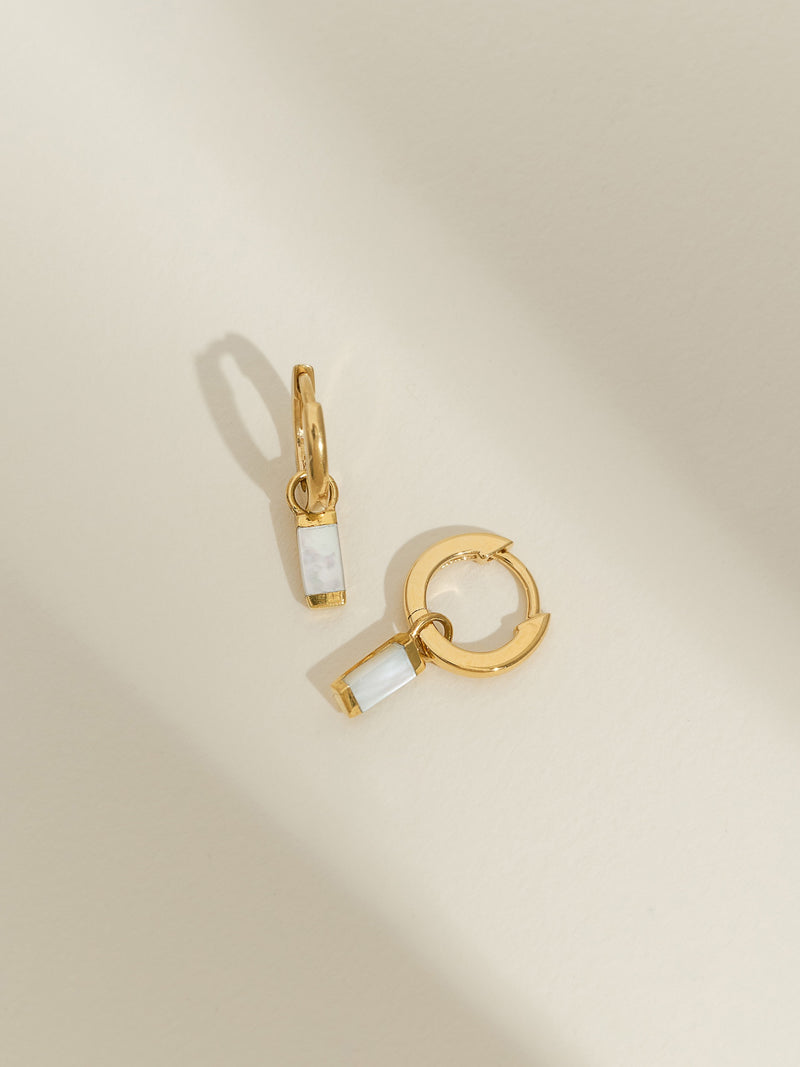 Gold-plated huggie hoop earrings with rectangular mother of pearl drop charms, displayed on a light cream background.