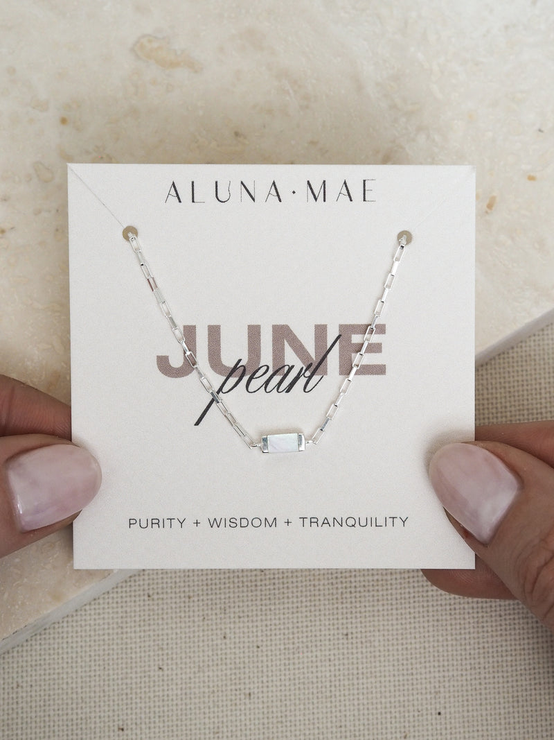 Hands holding a June birthstone bracelet featuring a rectangular mother of pearl stone on a sterling silver chain, displayed on a branded Aluna Mae card with the words 'Purity + Wisdom + Tranquility.
