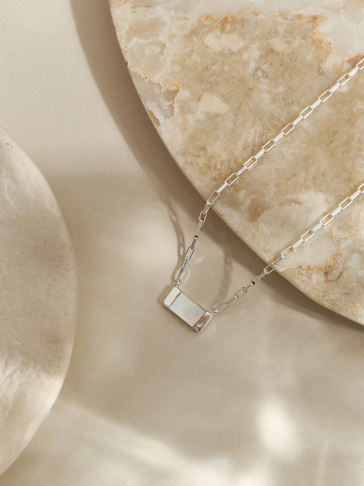 Sterling silver necklace featuring a rectangular mother of pearl pendant on a box chain, styled on a travertine stone surface with soft natural light.