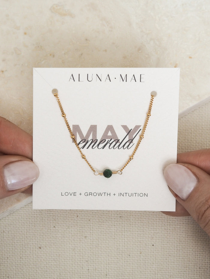 Close-up of hands holding a May birthstone necklace with a green emerald gemstone, on a delicate 18ct gold-plated sterling silver satellite chain, displayed on a card with the words 'MAY emerald' and the meanings 'Love + Growth + Intuition.