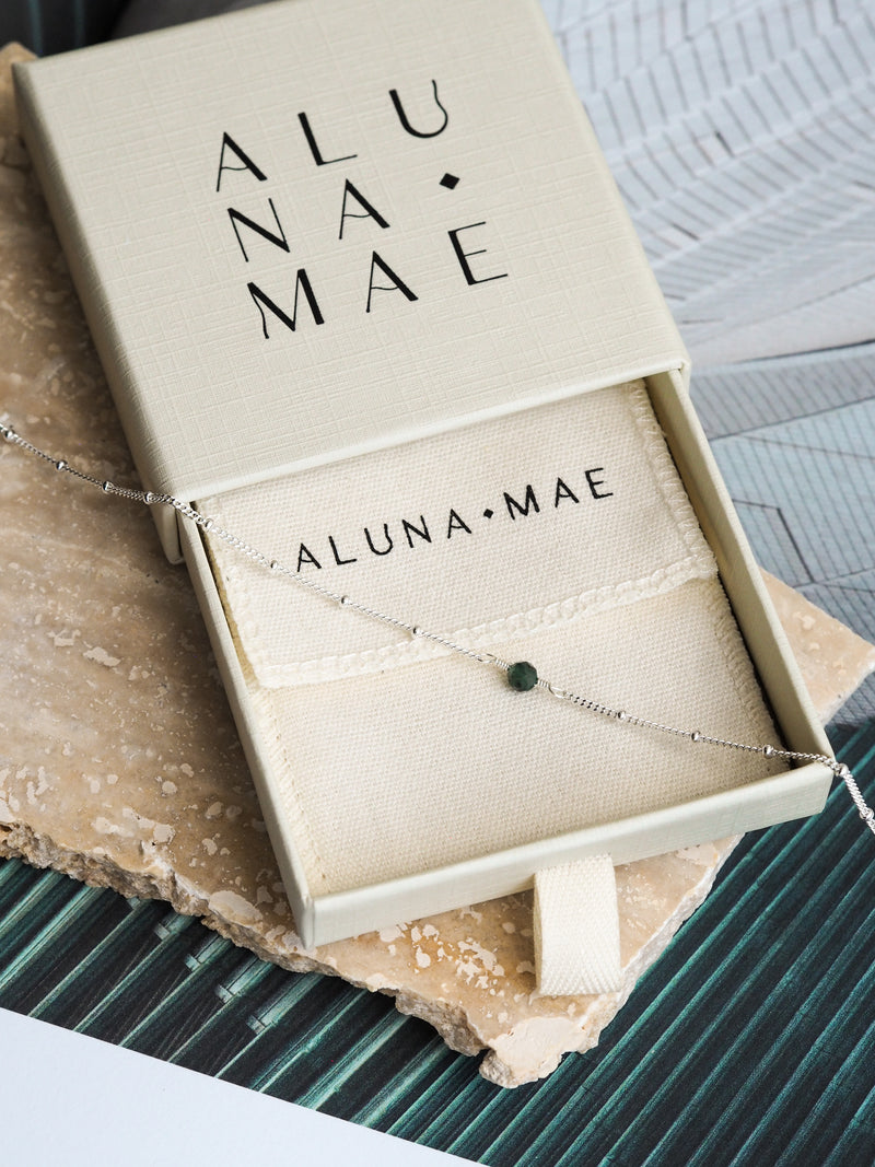 May birthstone necklace with a green emerald gemstone displayed in an Aluna Mae gift box, with the brand name visible on the interior fabric pouch and a sterling silver satellite chain.