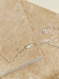 Sterling silver box chain bracelet with a rectangular mother of pearl pendant, styled on natural travertine stone with an Aluna Mae charm tag visible.