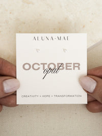 Close-up of hands holding a pair of October birthstone opal gemstone stud earrings, set in sterling silver, displayed on a card with the words 'October opal' and the meanings 'Creativity + Hope + Transformation.