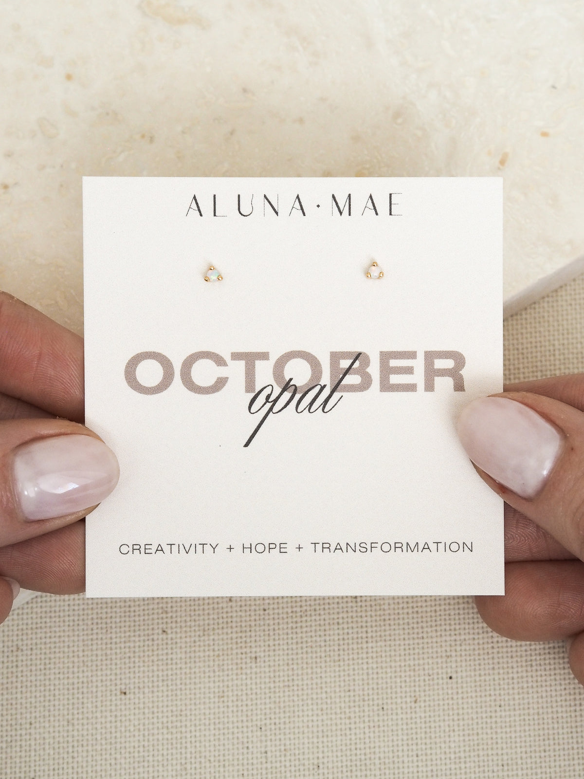 Close-up of hands holding a pair of October birthstone opal gemstone stud earrings, set in 18ct gold-plated sterling silver, displayed on a card with the words 'October opal' and the meanings 'Creativity + Hope + Transformation.