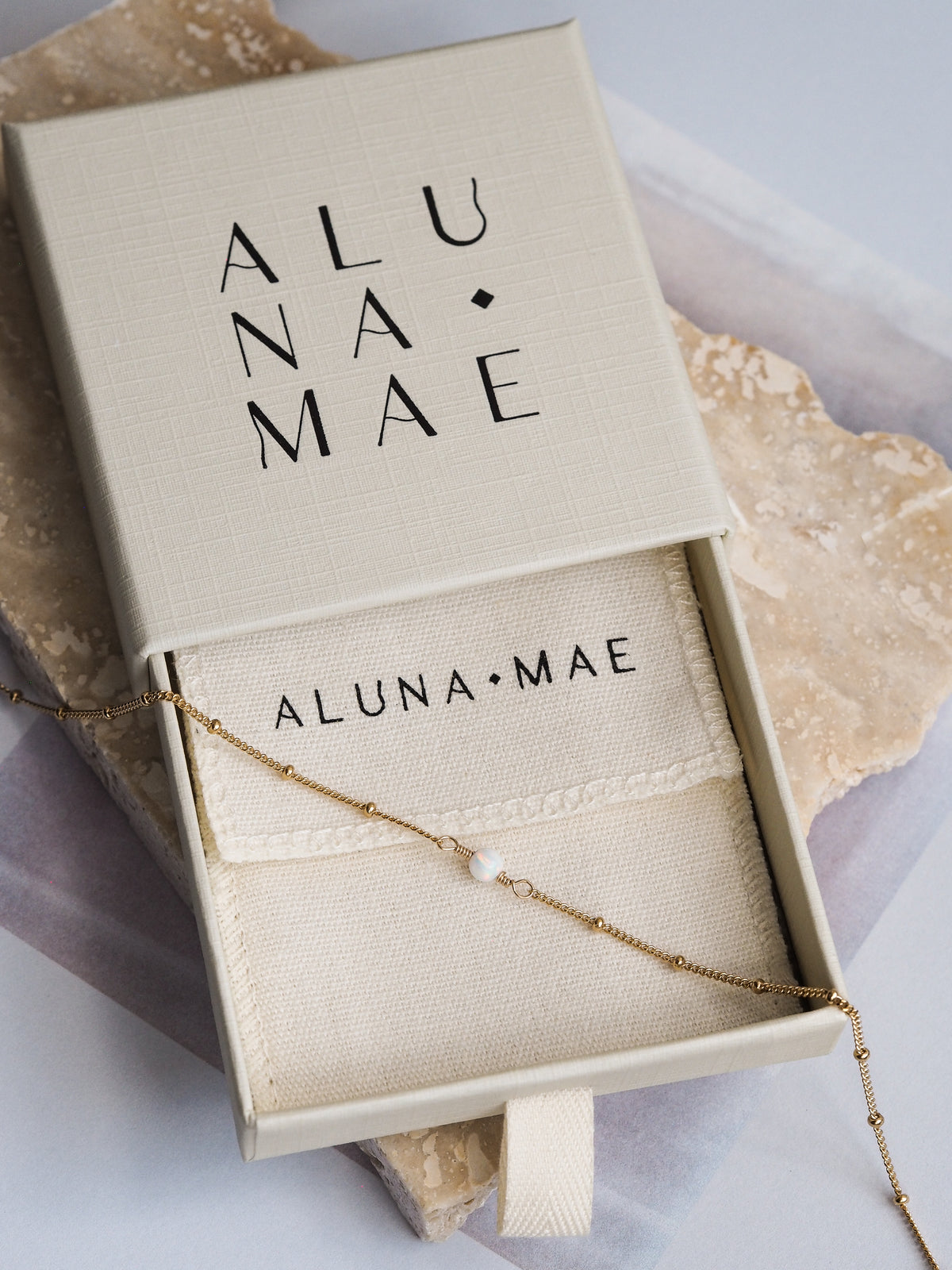 Aluna Mae necklace with a white opal gemstone displayed in a gift box, featuring a delicate 18ct gold-plated sterling silver chain. The brand name is visible on the interior fabric pouch.