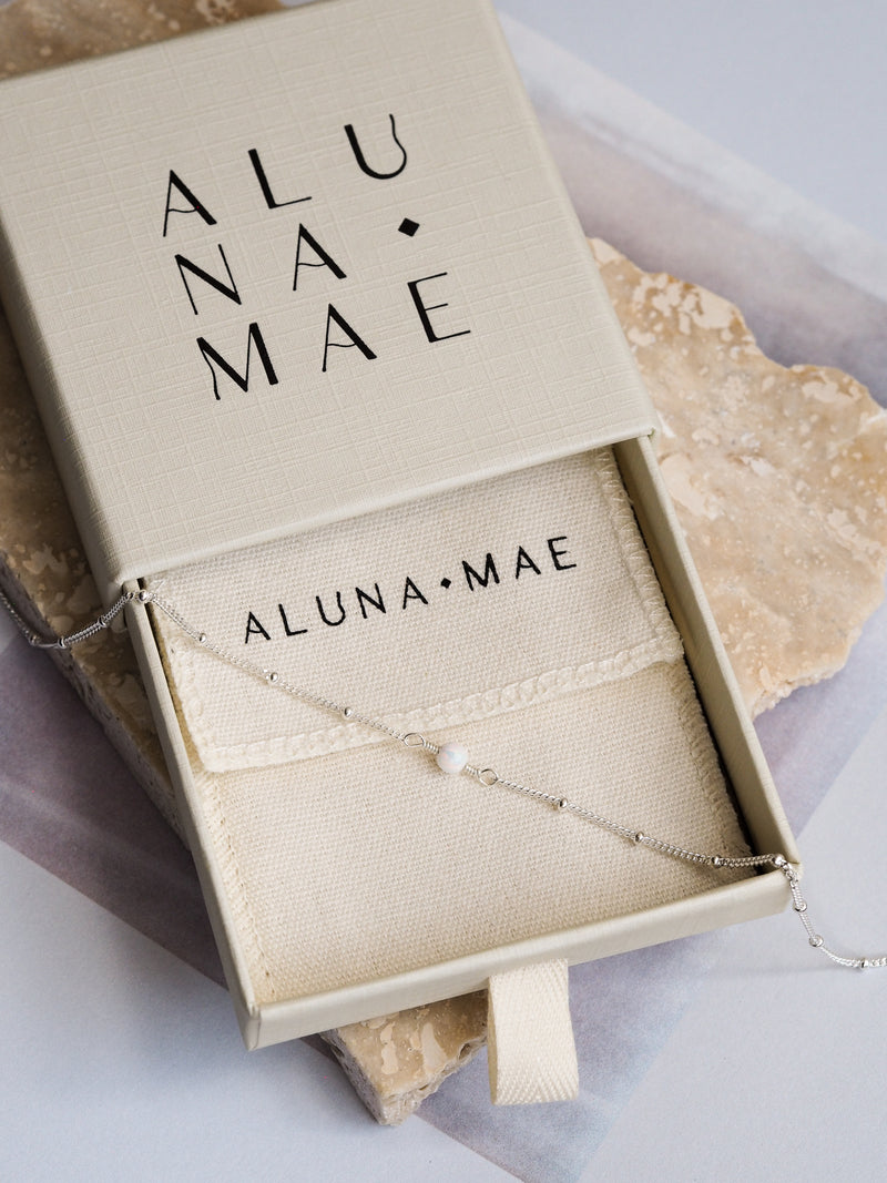 Aluna Mae necklace with a white opal gemstone on a sterling silver satellite chain, displayed in a gift box with the brand name visible.