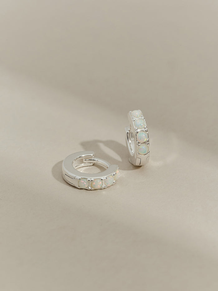 A pair of sterling silver huggie hoop earrings set with five round opal stones, displayed on a neutral-toned background.