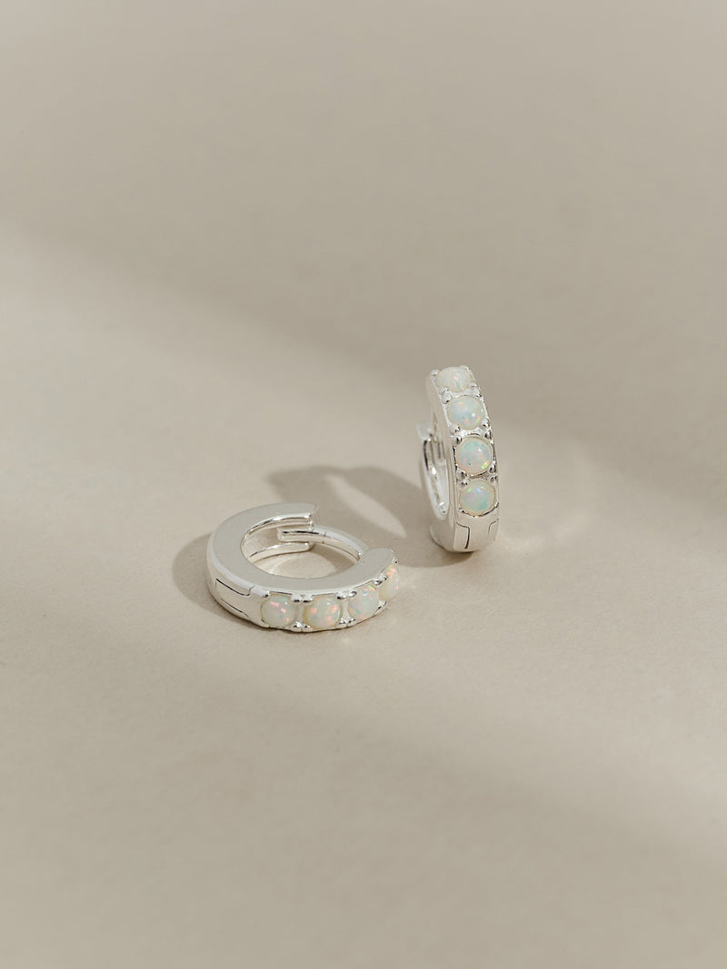A pair of sterling silver huggie hoop earrings set with five round opal stones, displayed on a neutral-toned background.