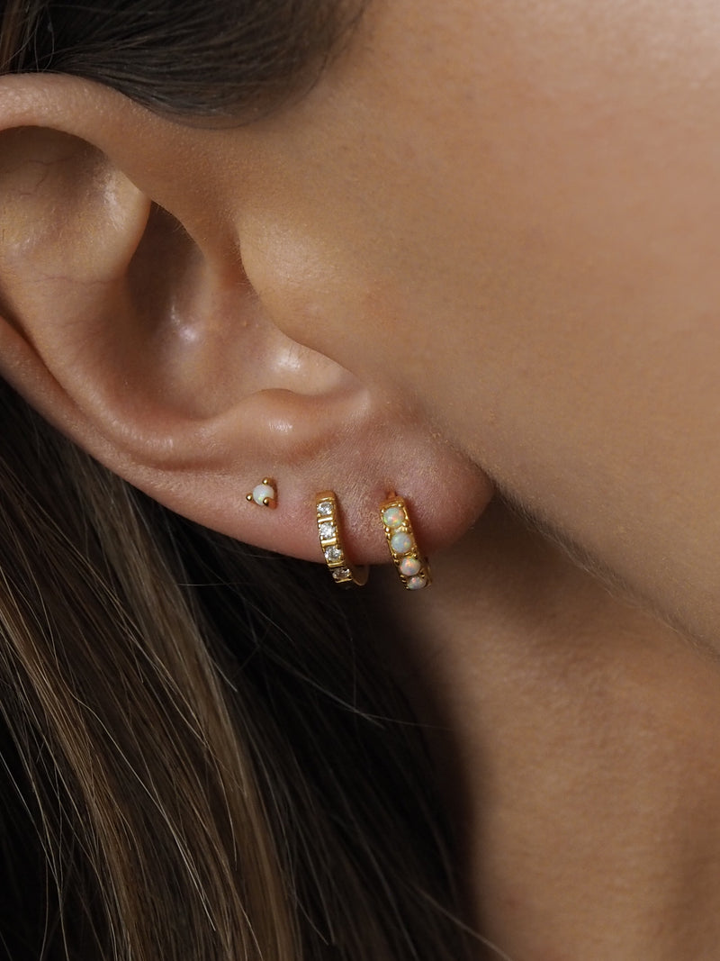 Close-up of three gold earrings worn in a lobe stack: a small opal stud and two gold huggie hoops set with opal and cubic zirconia stones.