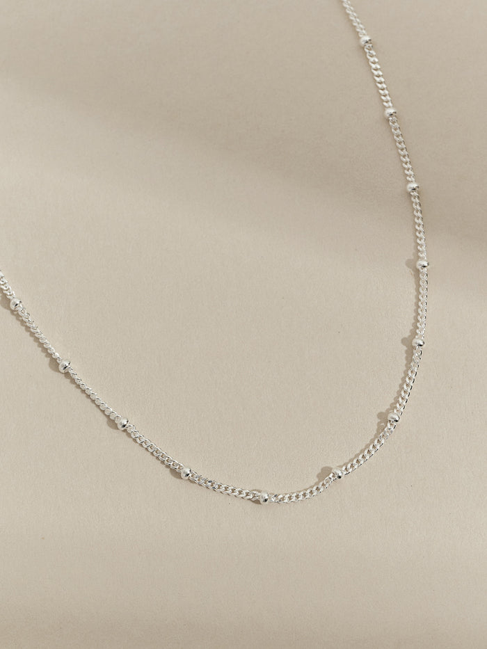 Close-up of a sterling silver satellite chain necklace with small evenly spaced beads, laid flat on a neutral background.