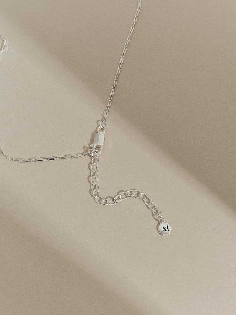 Close-up of a sterling silver extender chain with lobster clasp and 'AM' branded charm, styled on a soft neutral background.