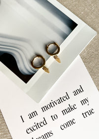 Close-up of gold hoops with a small diamond-shaped drop pendant, displayed on a polaroid and affirmation card.