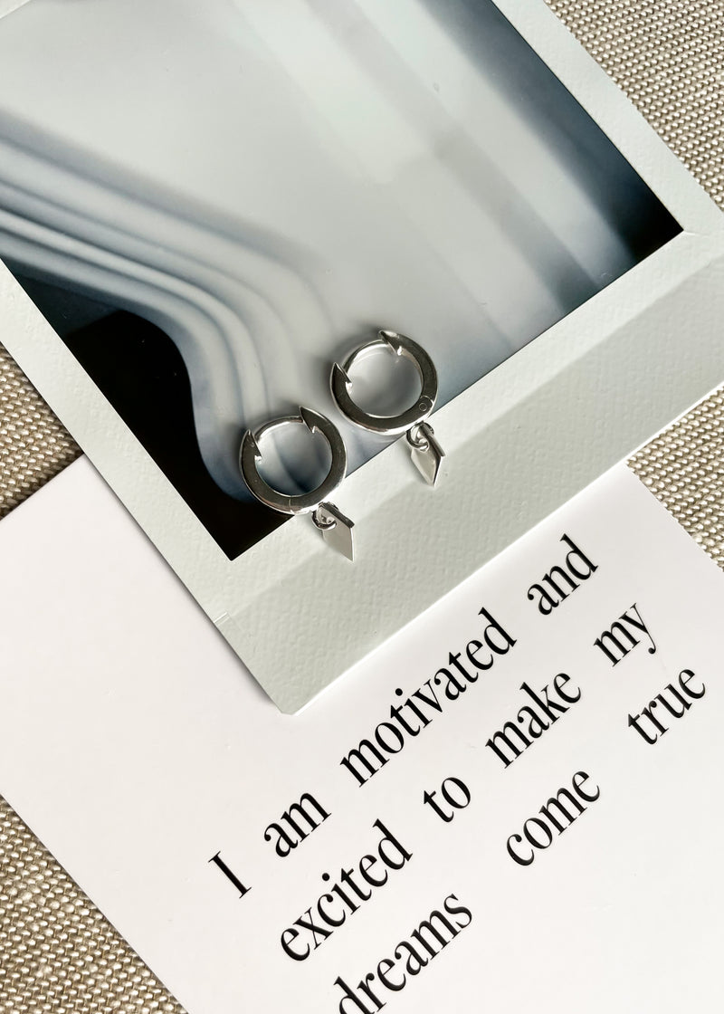 Close-up of sterling silver hoops with a small diamond-shaped drop pendant, displayed on a polaroid and affirmation card.