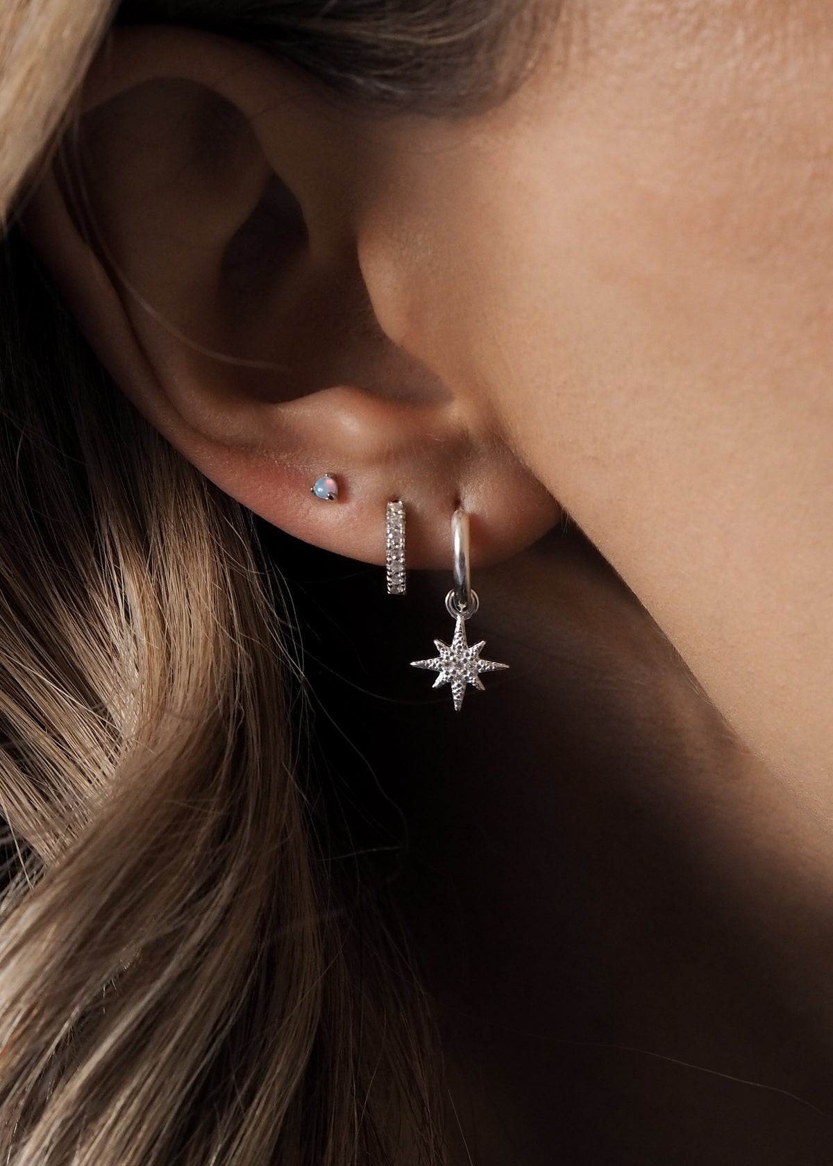  A model wearing multiple sterling silver earrings layered together, featuring star hoops, opal studs and huggie details.