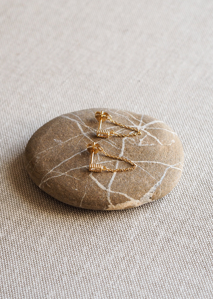 A pair of gold-plated ribbed stud earrings with delicate chain drops, styled on a smooth stone with white veining.