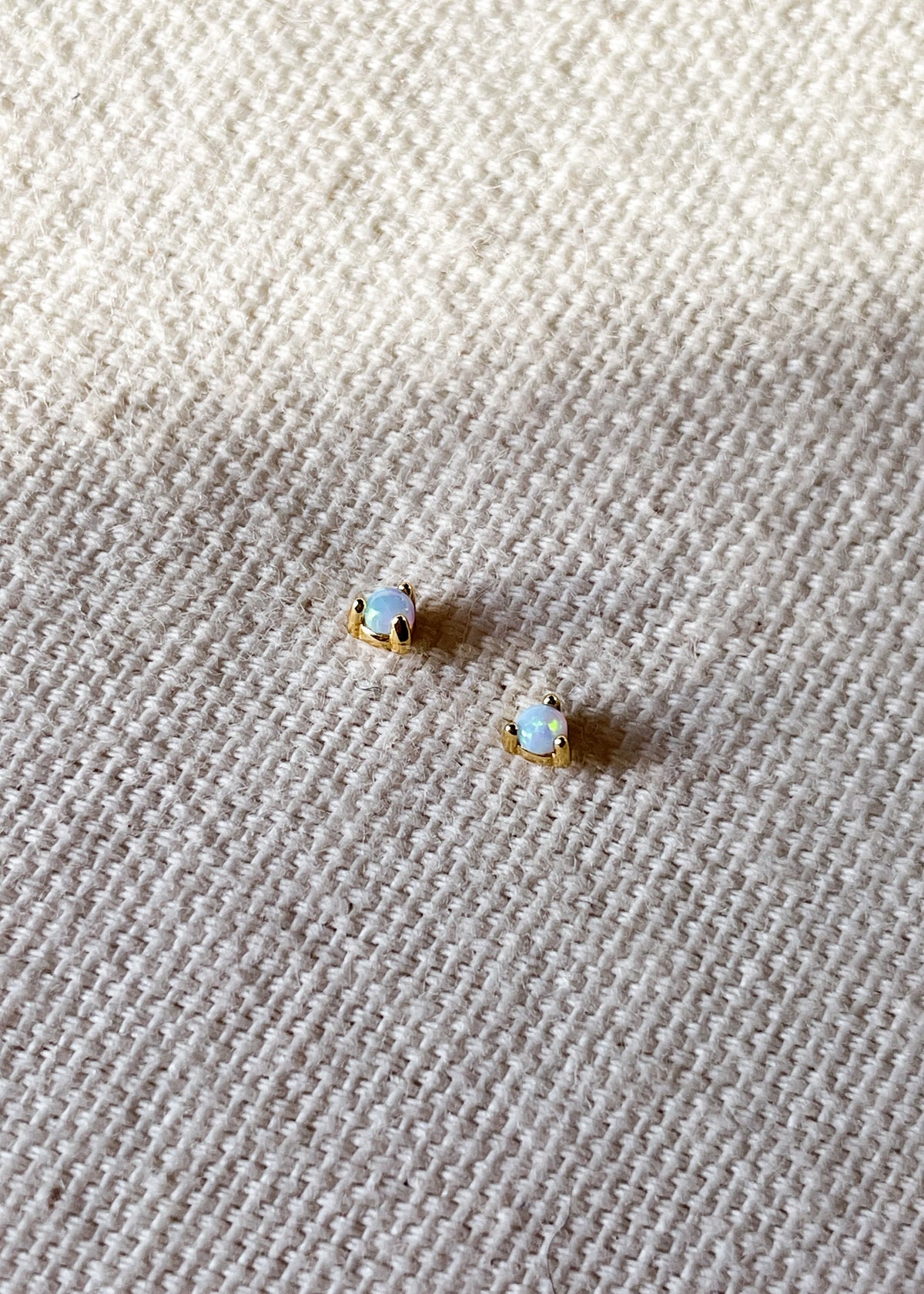 Pair of October birthstone opal gemstone stud earrings set in 18ct gold-plated sterling silver, displayed on a textured cream fabric background.