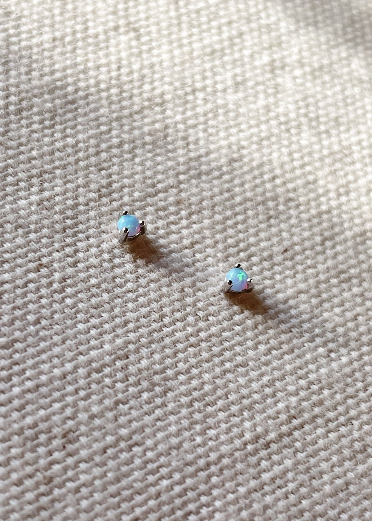 Pair of October birthstone opal gemstone stud earrings set in sterling silver, displayed on a textured cream fabric background.