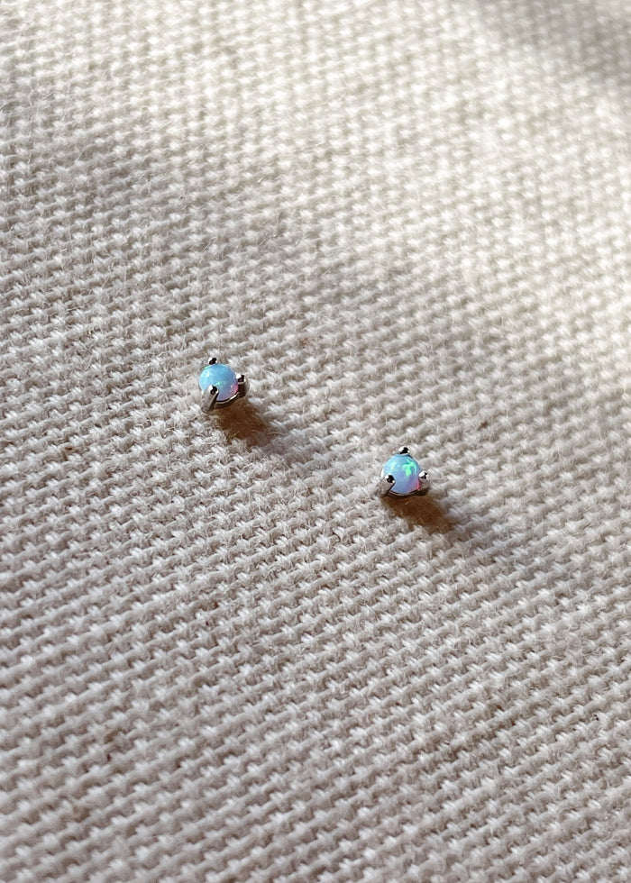Pair of October birthstone opal gemstone stud earrings set in sterling silver, displayed on a textured cream fabric background.