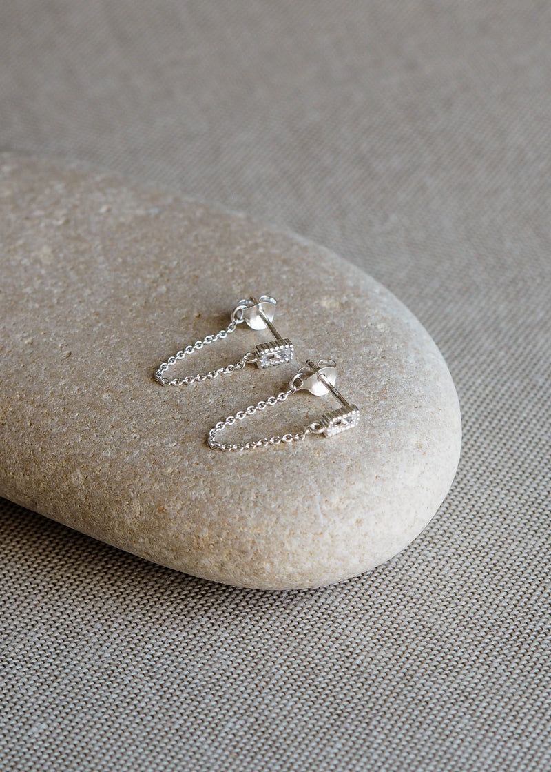 Pair of sterling silver drop chain earrings with rectangular cubic zirconia studs, displayed on a smooth neutral-toned stone.