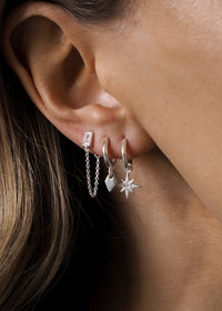 Three silver earrings worn on one ear: a rectangular cubic zirconia stud with drop chain, a mini tag charm hoop, and a starburst charm hoop.